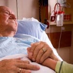Old man lying in hospital bed holding daugher's hand