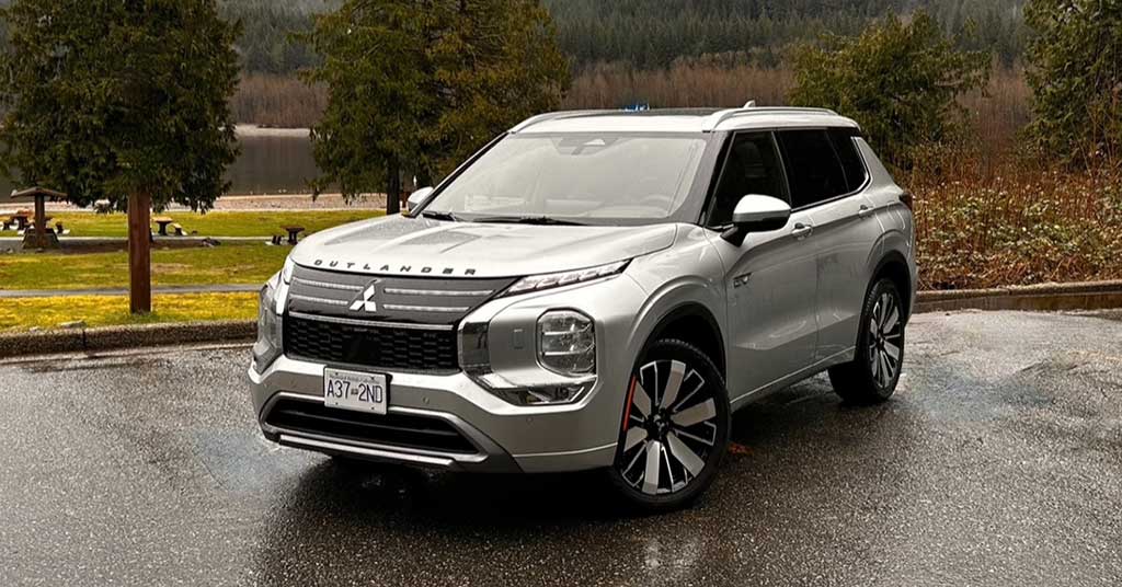 2026-Outlander-PHEV - The Bluenose Bulletin Front-three-quarter view of a silver Mitsubishi Outlander SUV parked on a wet pavement with trees and a lake in the background, daytime outdoor scene.