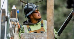 Construction worker in a hard hat and safety glasses looks up at equipment on a construction site with a perforated metal panel nearby.